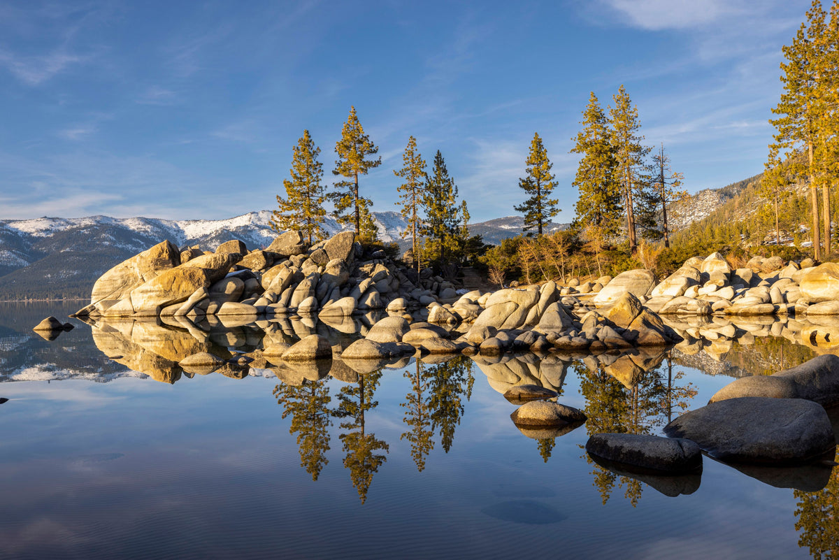 Calm clear lake reflecting granite boulders and pine trees under a blue sky